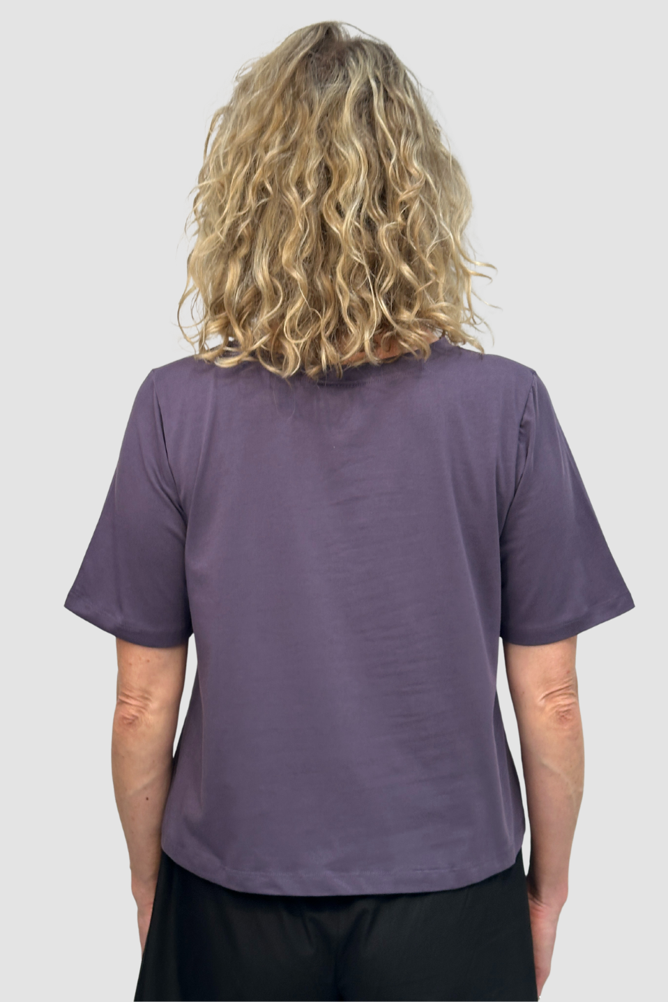 Back view of woman with curly blonde hair wearing a loose purple short-sleeve shirt and black pants