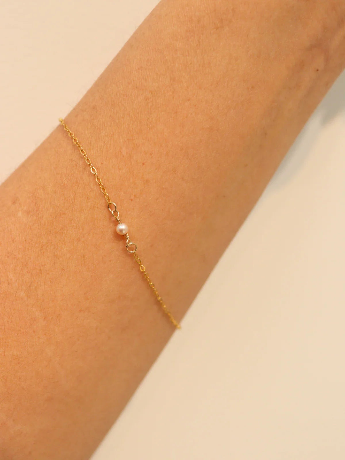 Gold bracelet with a single pearl on a wrist against a beige background