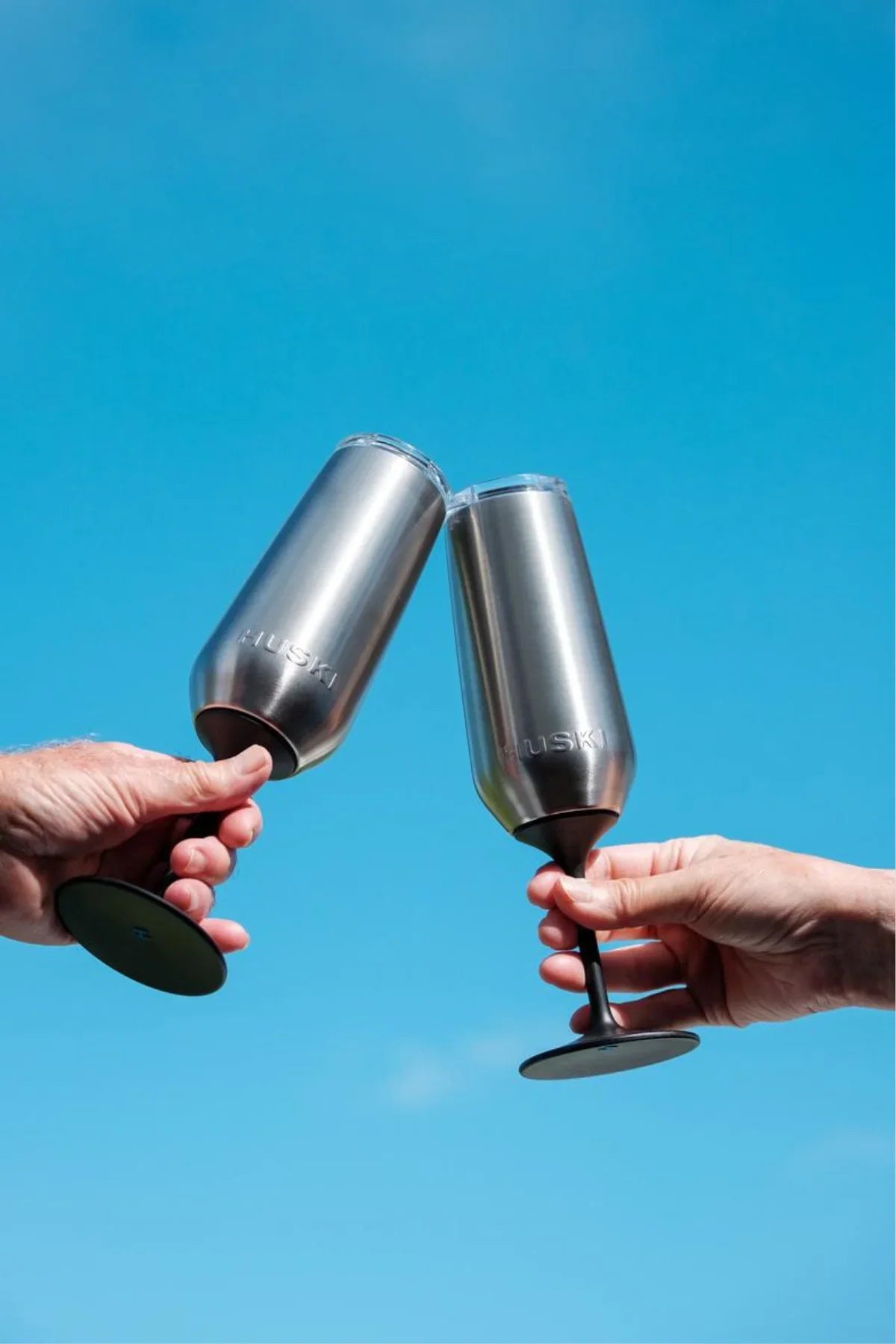 Two hands holding metallic wine glasses against a clear blue sky