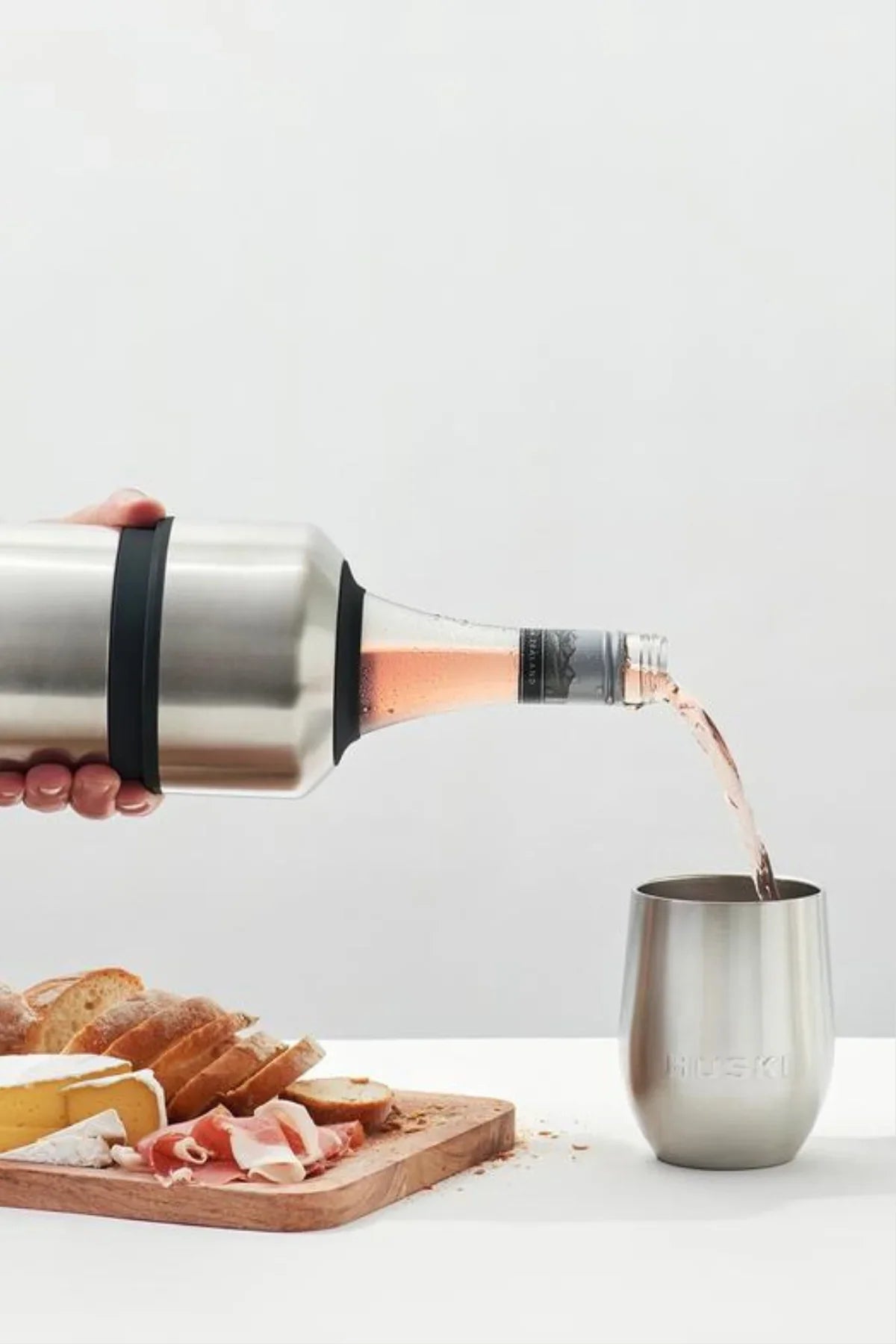 Person pouring rose wine from a bottle into a stainless steel tumbler with bread and prosciutto on a cutting board.