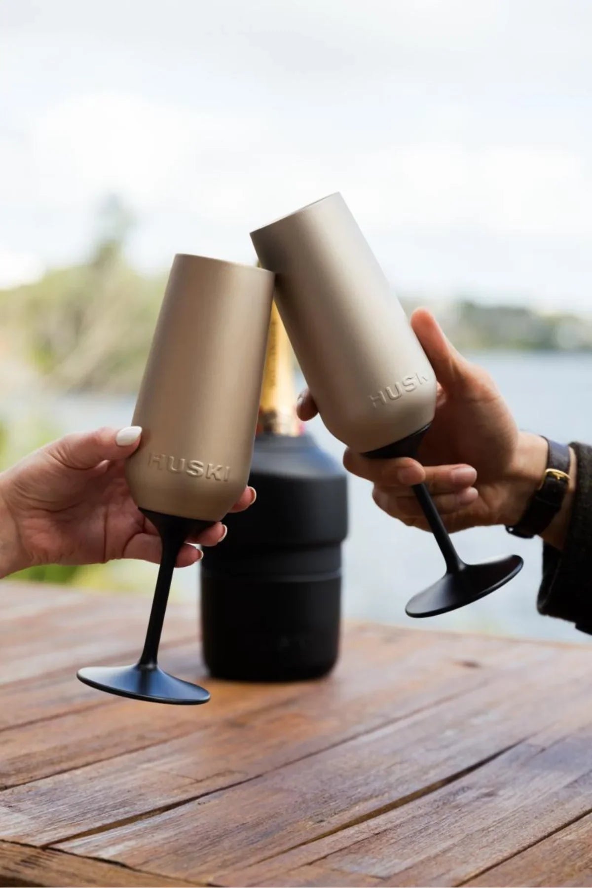 Two hands clinking Huski reusable cups against a blurred outdoor background.
