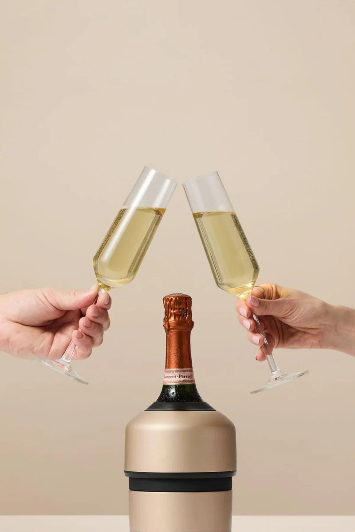 Two hands clinking champagne glasses with a bottle of champagne between them against a beige background