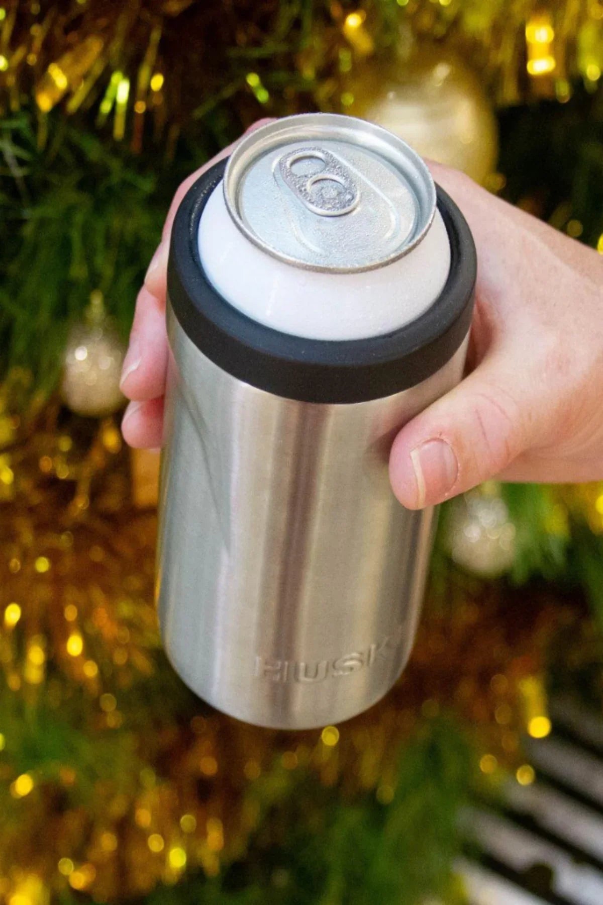 Hand holding a stainless steel can cooler with a Christmas tree in the background