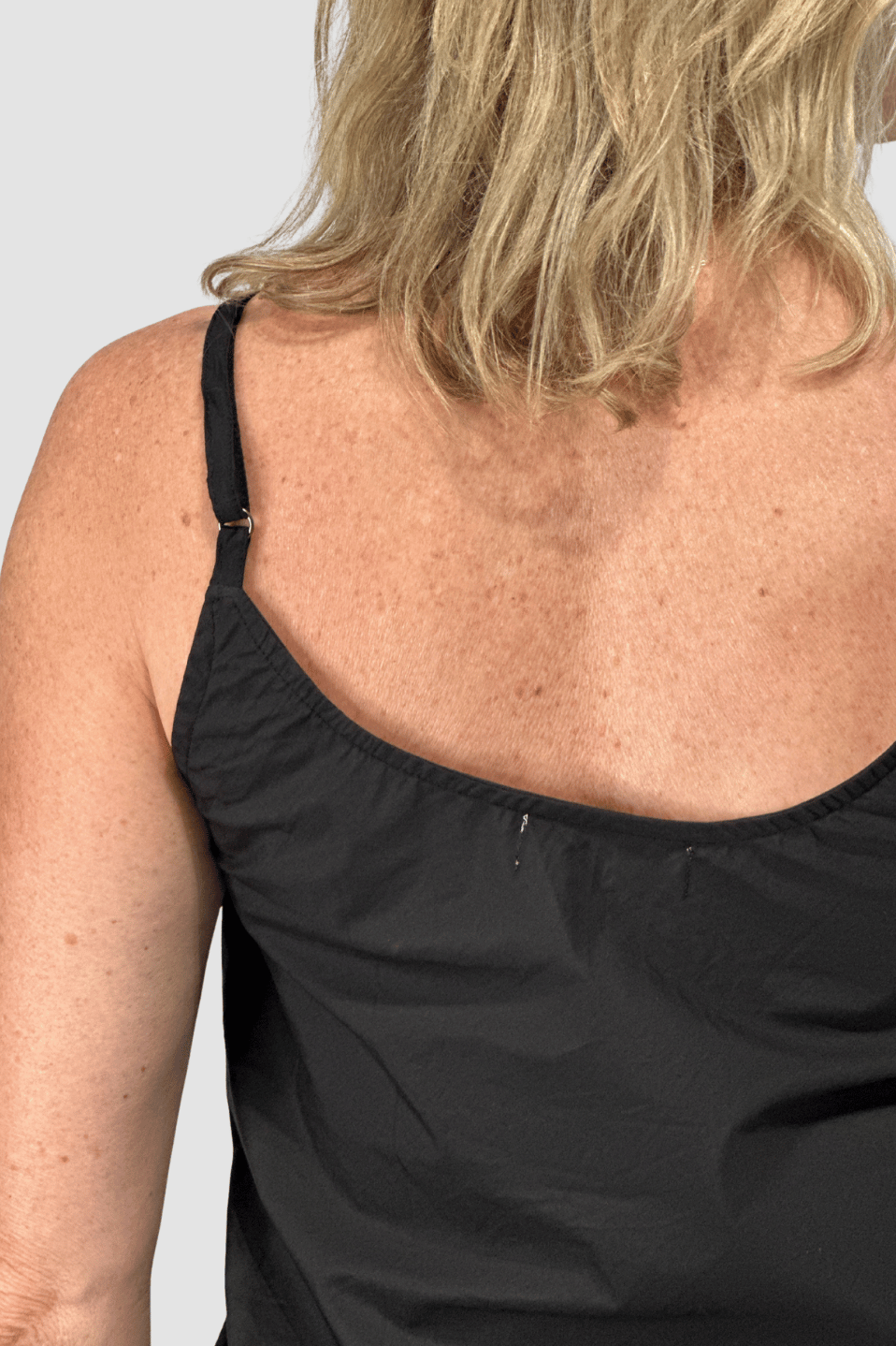 Close-up of a woman’s back wearing a black sleeveless ethical clothing top with adjustable strap, fair skin and blonde hair