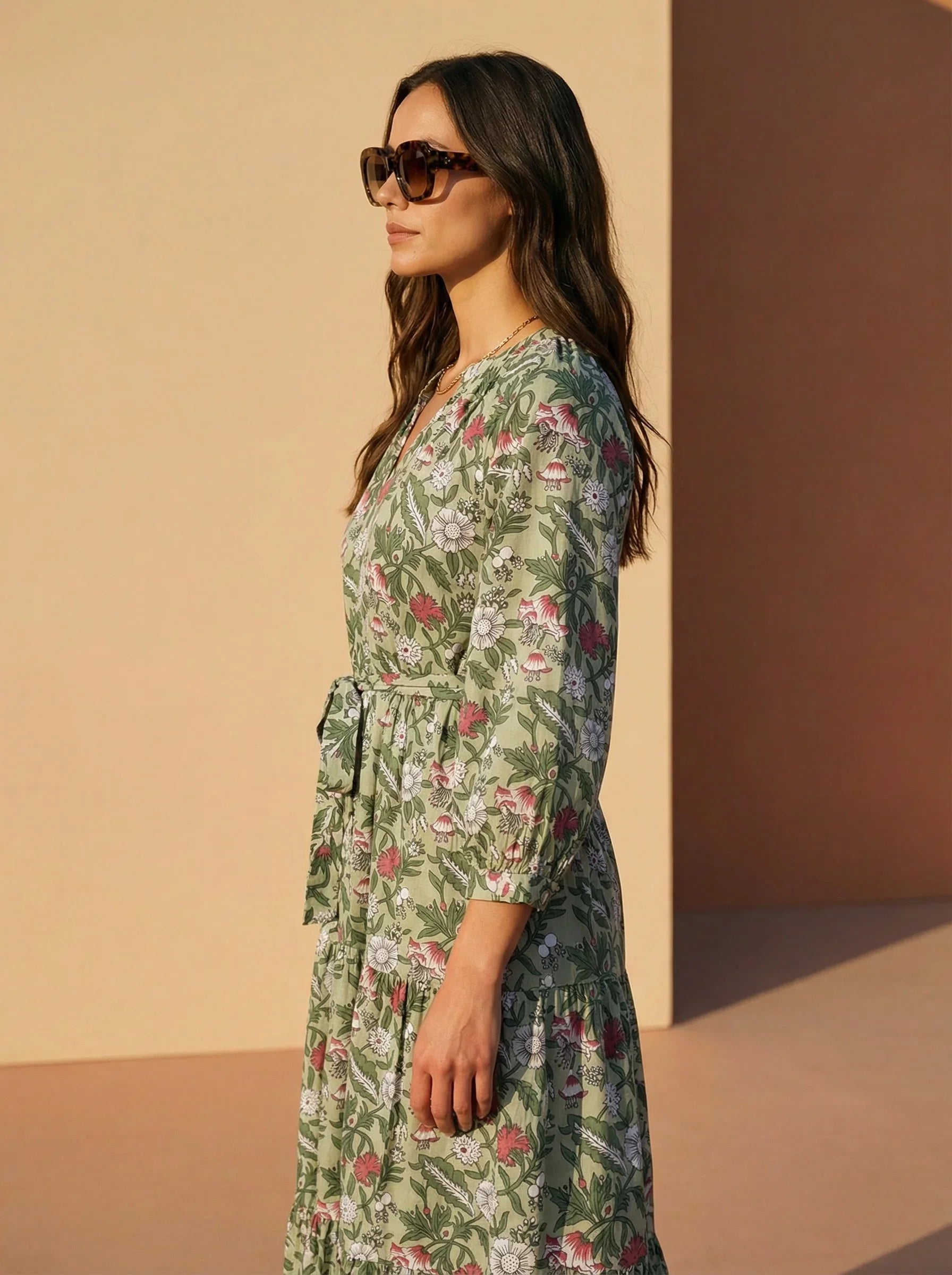 Woman wearing an ethical Reca gold necklace with THE CAMILLE DRESS – GREEN FLORAL. The model stands in a side profile pose, showing the side silhouette of the dress and the soft movement of the fabric. Her head is slightly turned toward the camera so the Reca gold necklace is visible. Clean, simple background with soft natural light. Minimalist fashion editorial style.