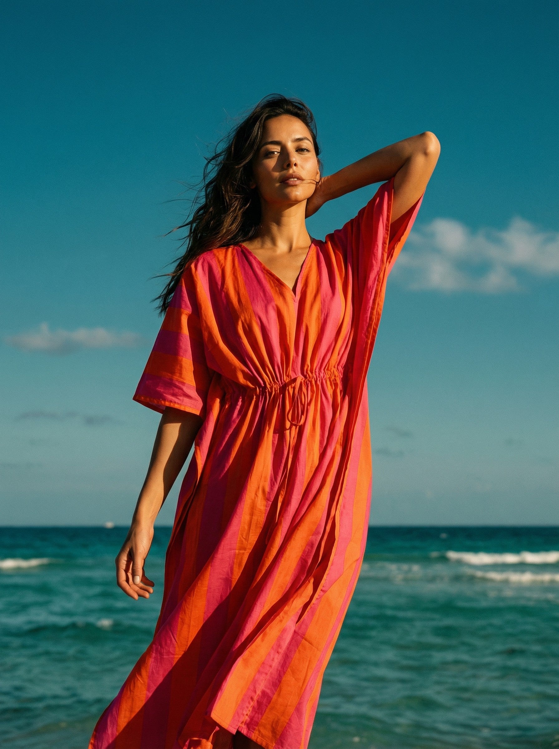 Woman in a bright orange dress standing on a beach with blue sky and ocean.