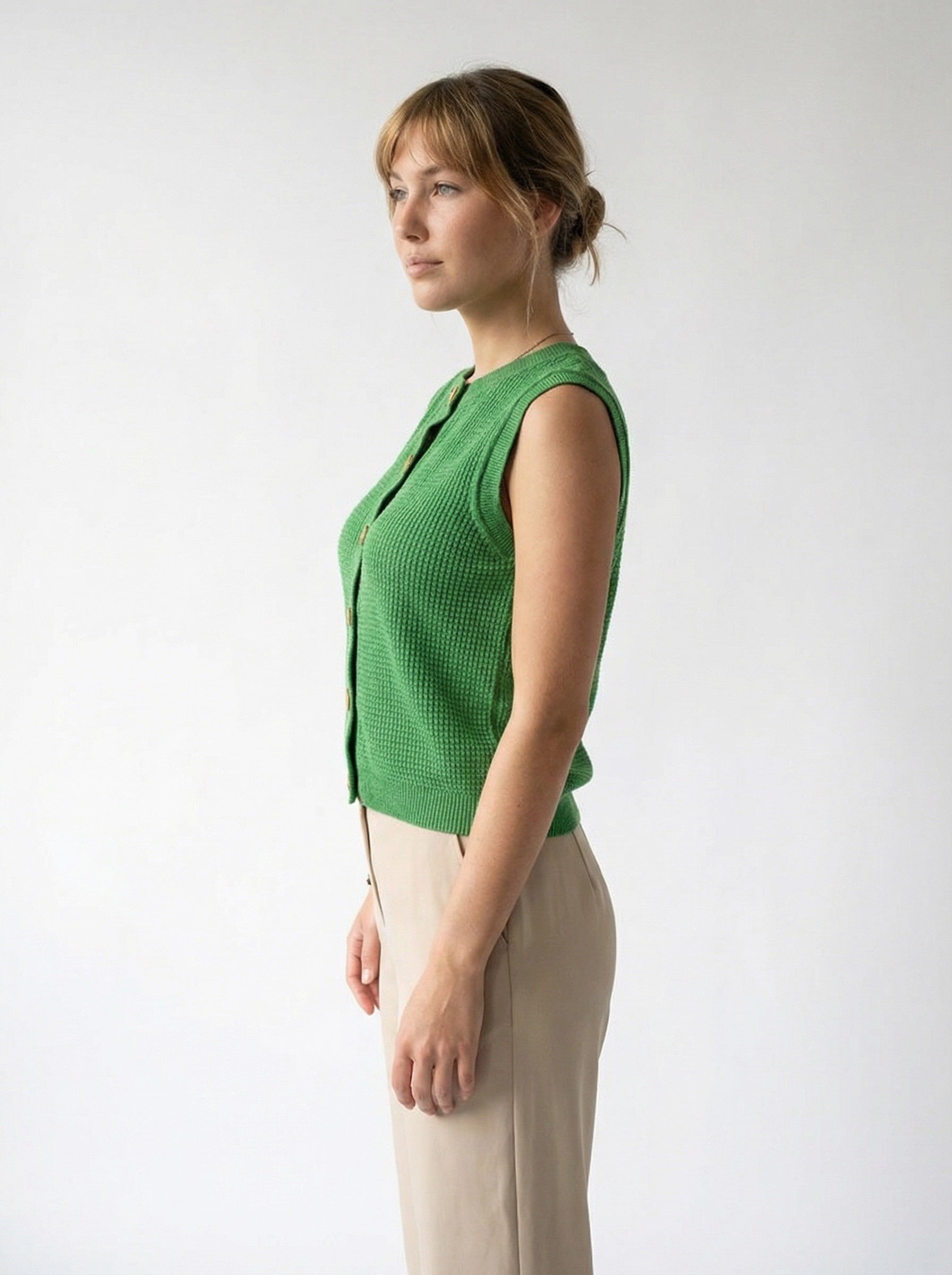 Woman wearing a COTTON & MERINO HEPBURN BUTTON VEST - KELLY GREEN and beige pants against a white background