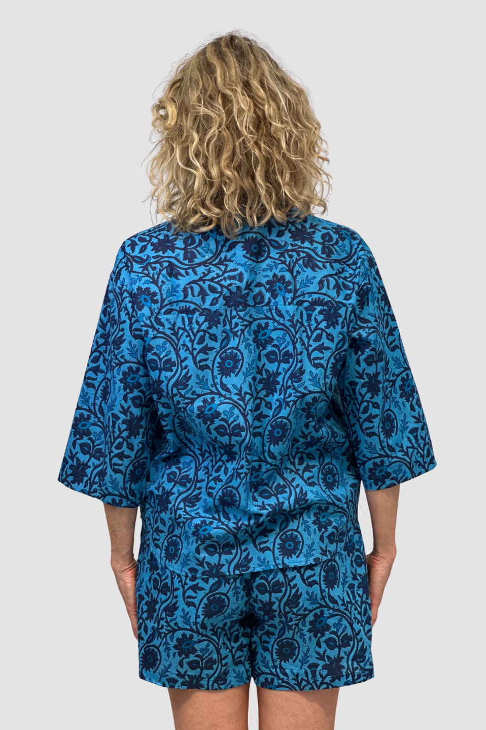 Woman with curly blonde hair wearing a blue floral patterned top, Named the "Indigo Tide Button Down" and matching shorts, back view