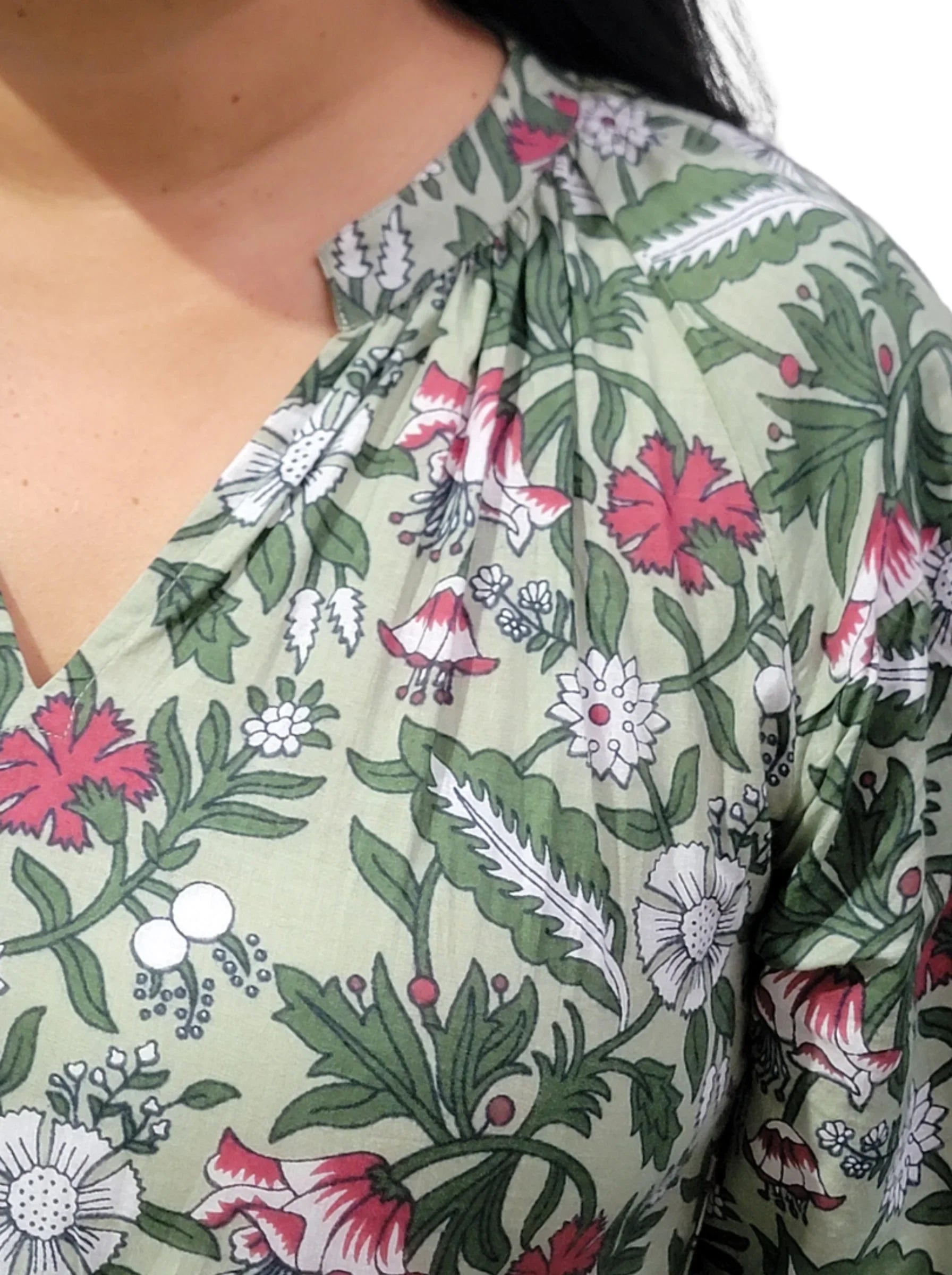 Close-up fashion editorial image of a woman wearing an ethical Reca gold necklace with THE CAMILLE DRESS – GREEN FLORAL. The frame focuses on the neckline, necklace, and upper bodice of the dress, highlighting the V-neckline and the floral print detail. The Reca gold necklace sits elegantly against the skin and is the main focal point. Soft natural light, clean simple background, minimalist and refined aesthetic, high-end fashion campaign style.