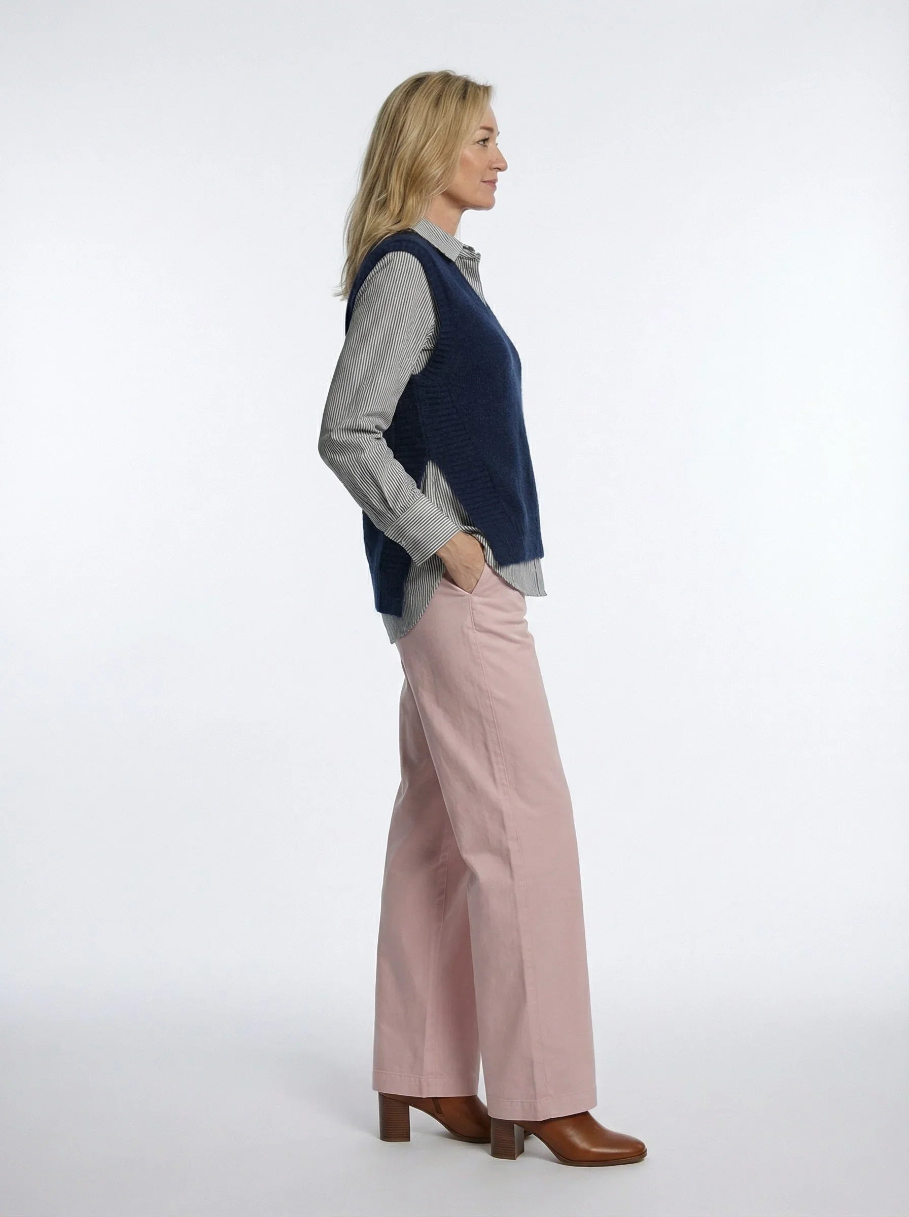 Woman in striped button-up shirt, navy vest, pink wide-leg pants and brown boots, side view