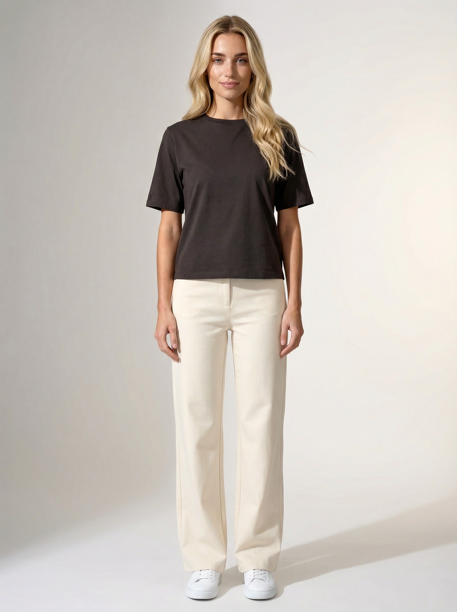 Woman wearing a black short-sleeve t-shirt, cream straight-leg pants, and white sneakers in a studio