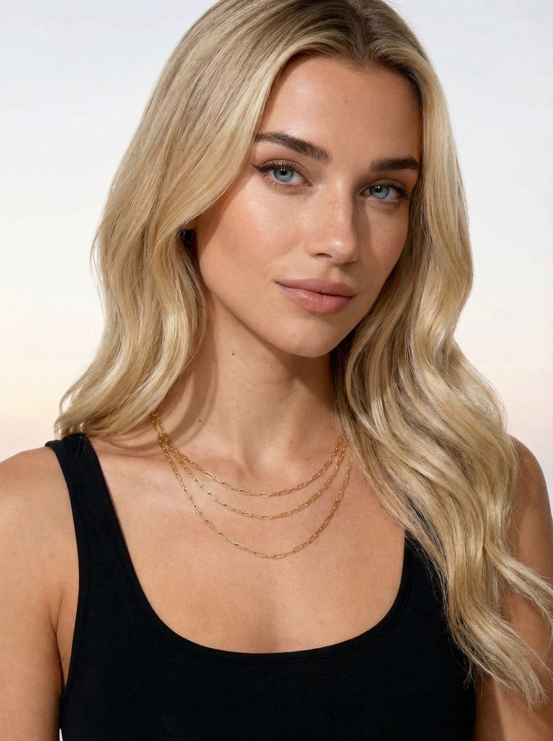 Woman with long blonde hair wearing a black top and gold filled paper clip chain necklace on a neutral background