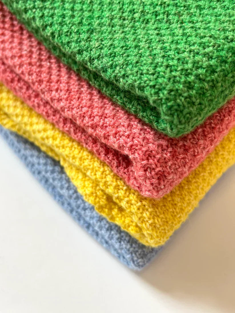 Stack of folded Cotton and Merino Hepburn Button Vests in green, pink, yellow and blue colours showing textured knit