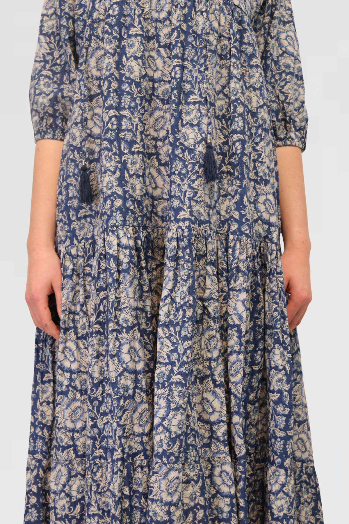 Close-up of Serene Petals Maxi Dress in blue floral cotton, showing V-neckline with tassel ties and tiered skirt detail.