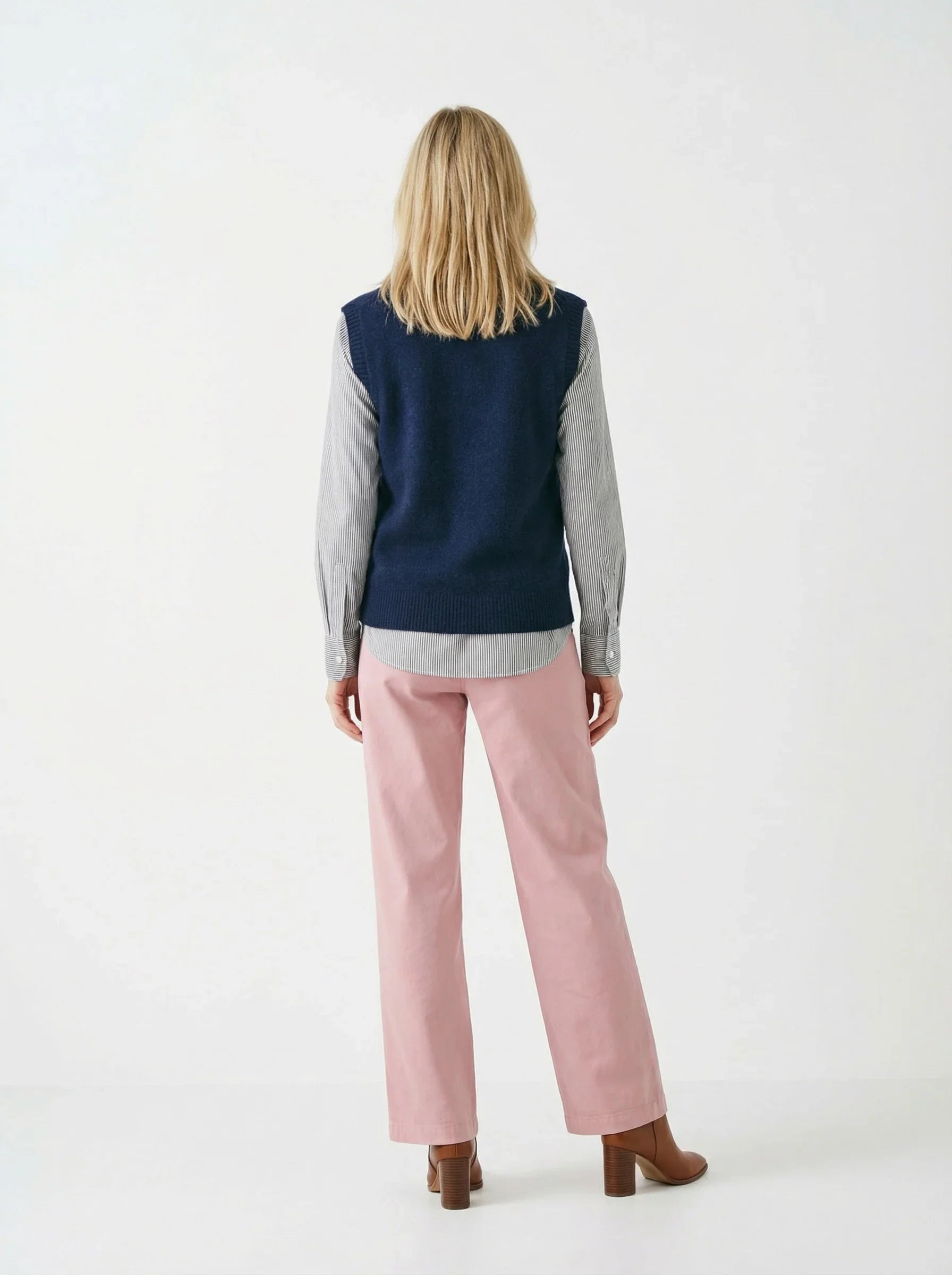 Woman wearing a navy sweater vest, striped shirt, pink pants, and brown boots, back view