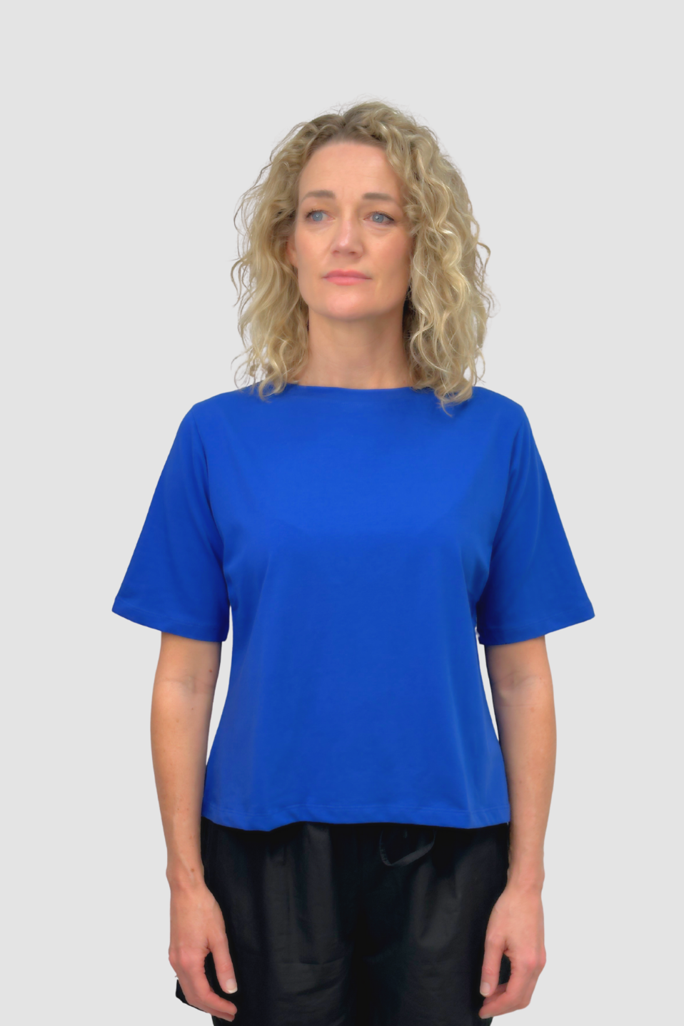 Woman with curly blonde hair wearing a plain blue short-sleeve t-shirt and black pants on gray background