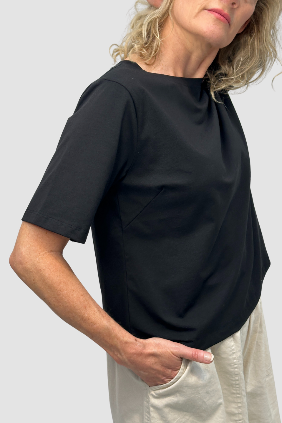 Woman wearing a black short-sleeve top and beige pants with hand in pocket on light gray background