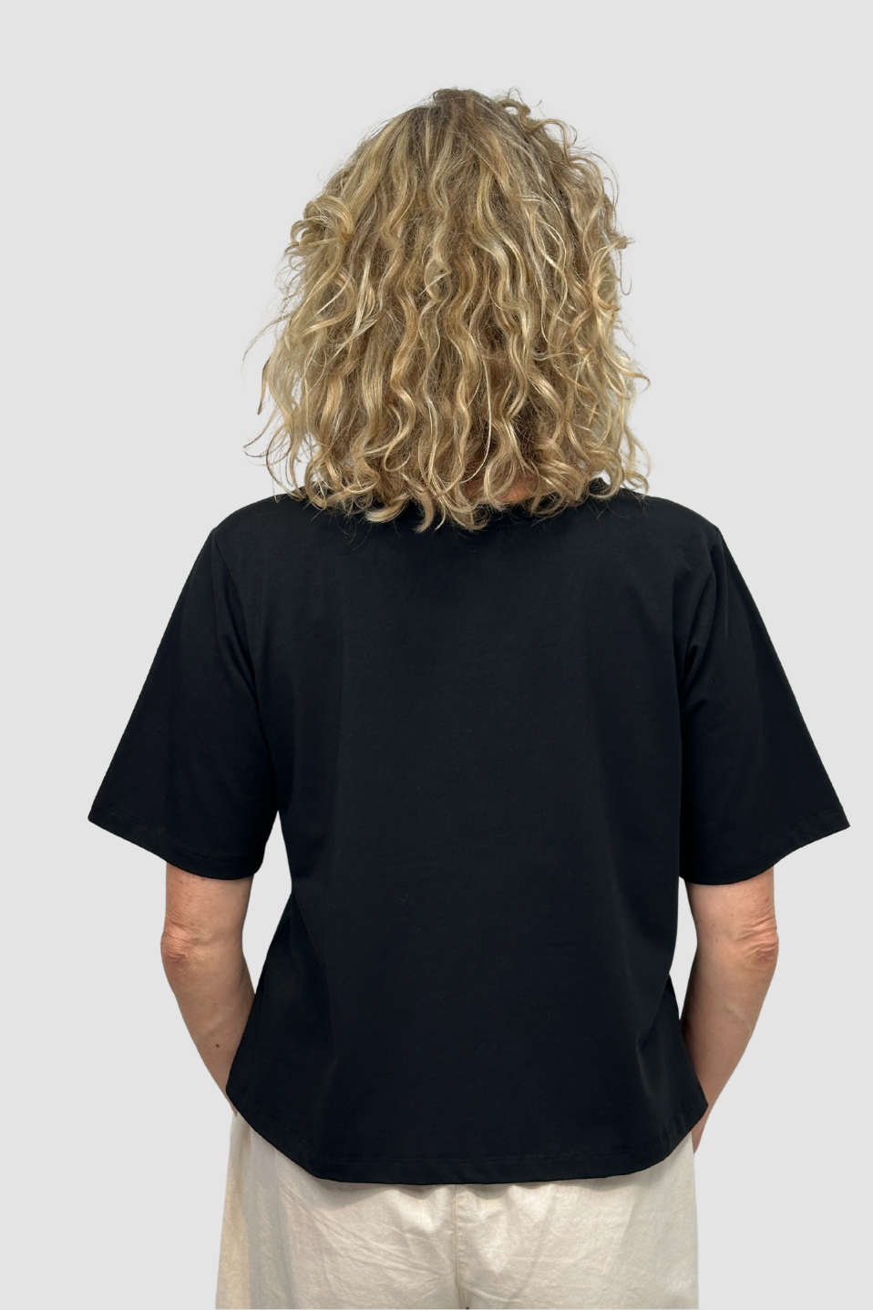 Back view of person with curly blonde hair wearing a black ethical cotton t-shirt and light pants