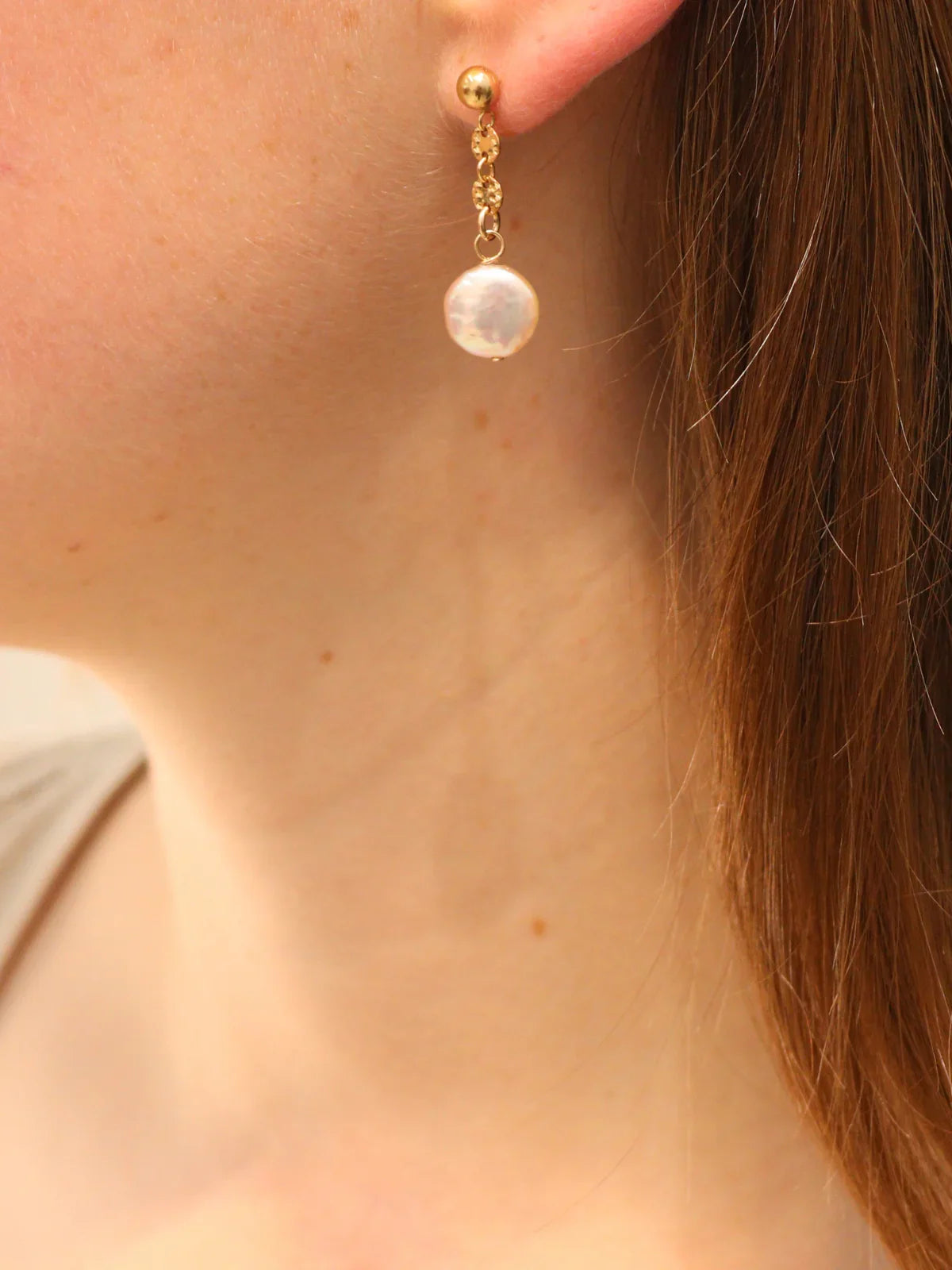 Delicate gold earring with dangling white pearl, shown on ear.