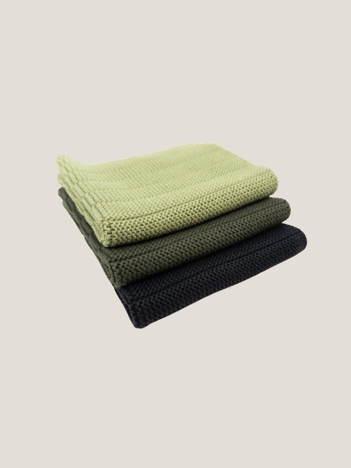 Stack of three folded knit dishcloth in green, olive, and black on a beige background