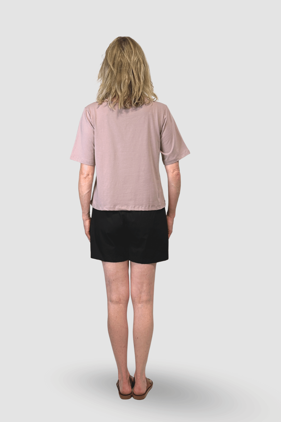 Back view of woman wearing a light mauve short-sleeve top, black shorts, and sandals on gray background