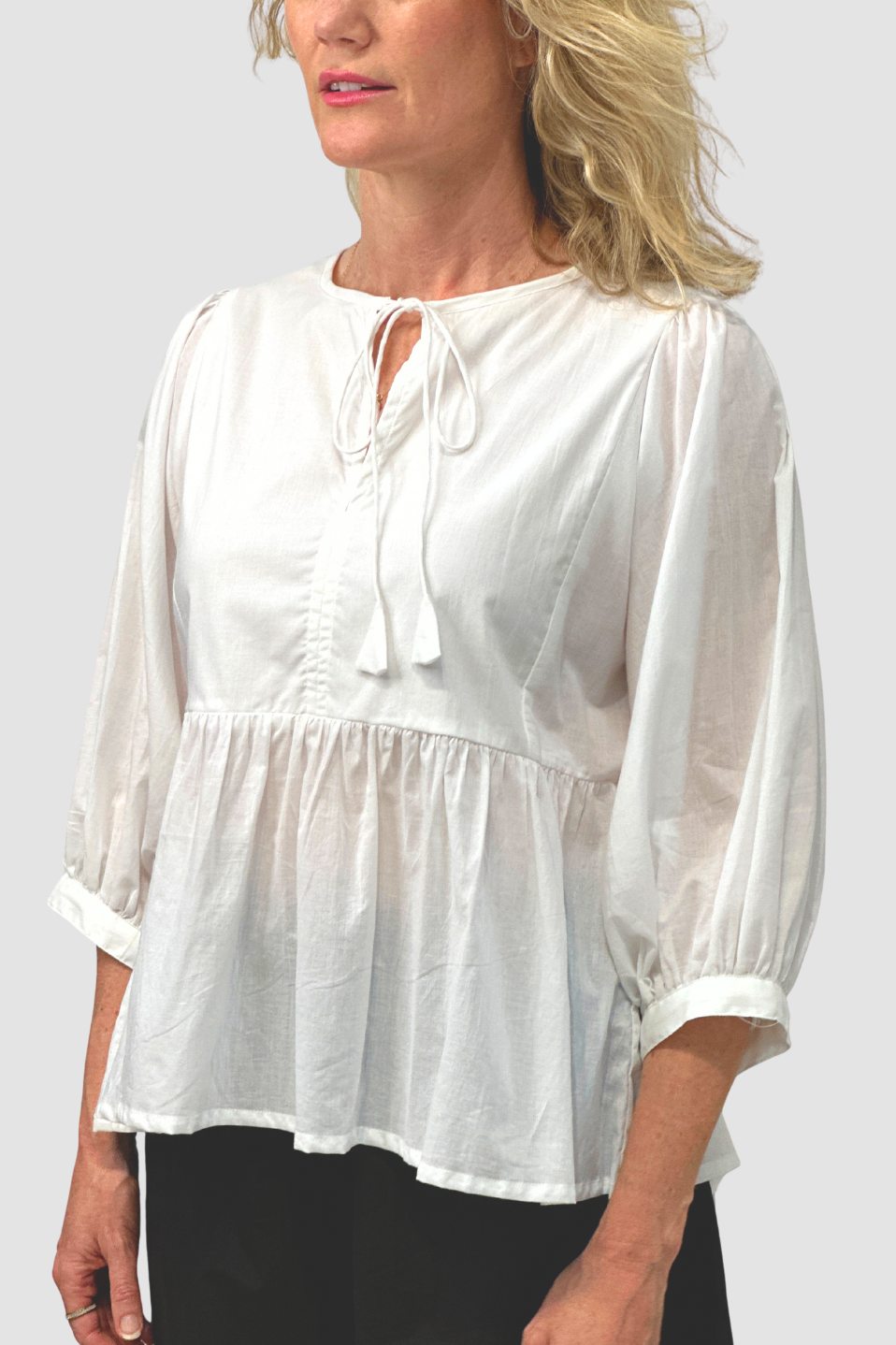 Woman wearing white loose cotton blouse with 3/4 puff sleeves and tie neckline on grey background