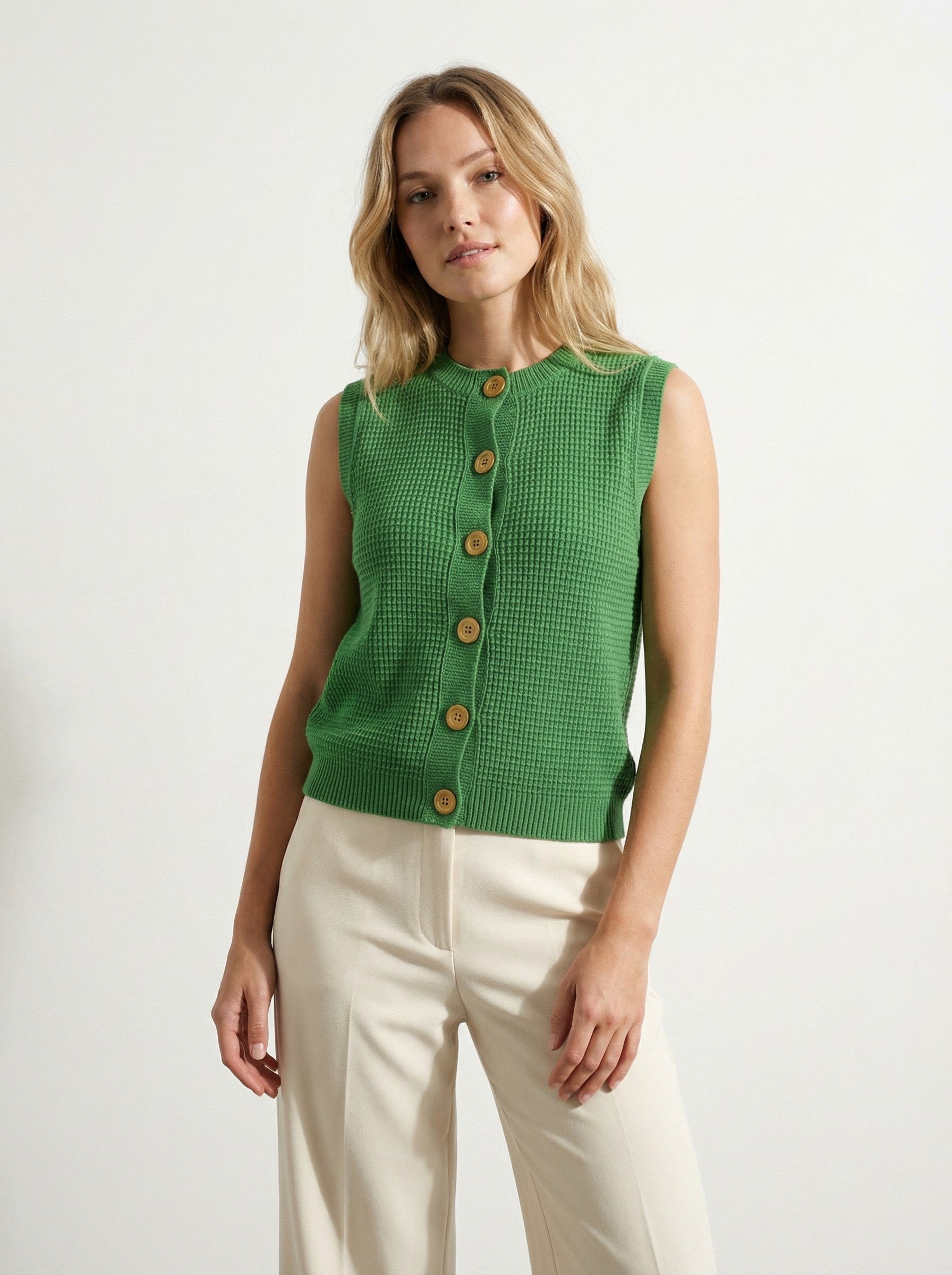 Woman wearing a COTTON & MERINO HEPBURN BUTTON VEST - KELLY GREEN with beige pants on a white background