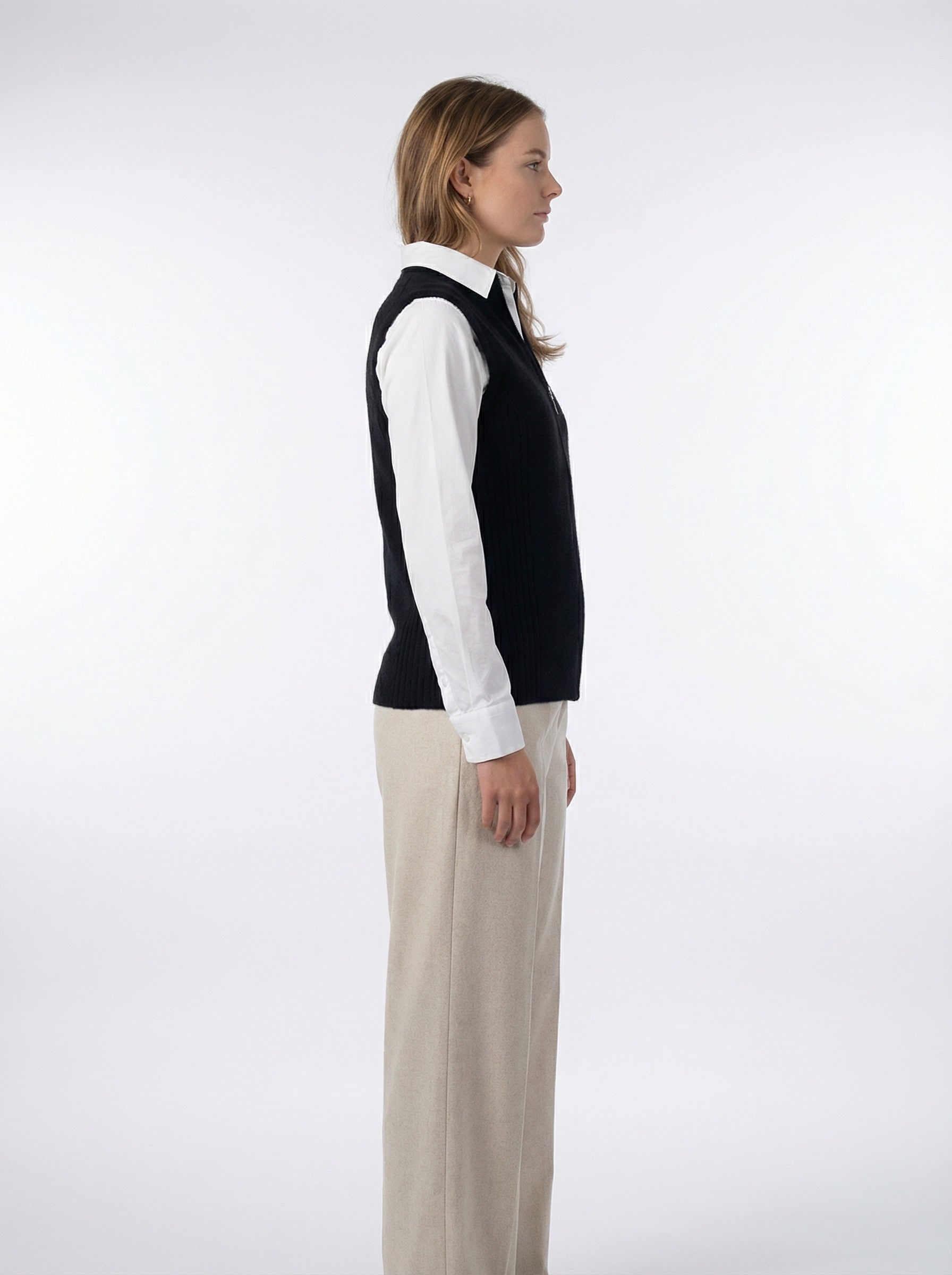 Woman wearing a Rib Zip Vest – Black over a white shirt with beige pants on a white background