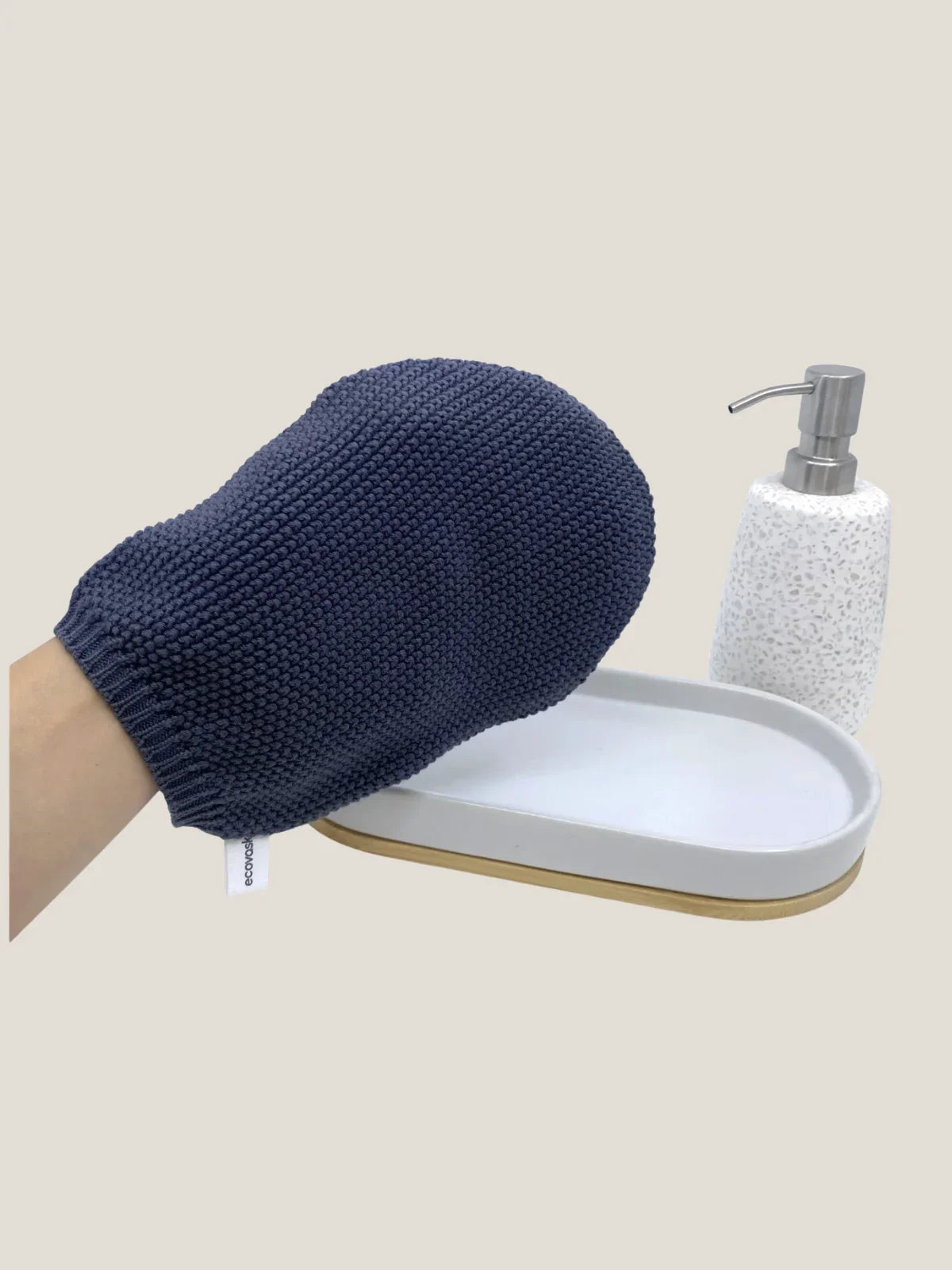 Blue exfoliating glove held over a white soap dish with a soap dispenser on a beige background