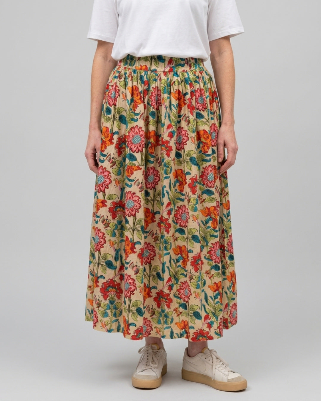 Woman wearing the Reca Bloomfield Garden Skirt, a cotton floral midi skirt in red, coral, orange and teal on a cream base, styled with a white tee and cream sneakers