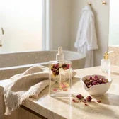 Nude Kiwi Body Oil Safflower & Rose bottle with dried rose petals on a marble bathtub ledge – luxury bath self-care lifestyle setting