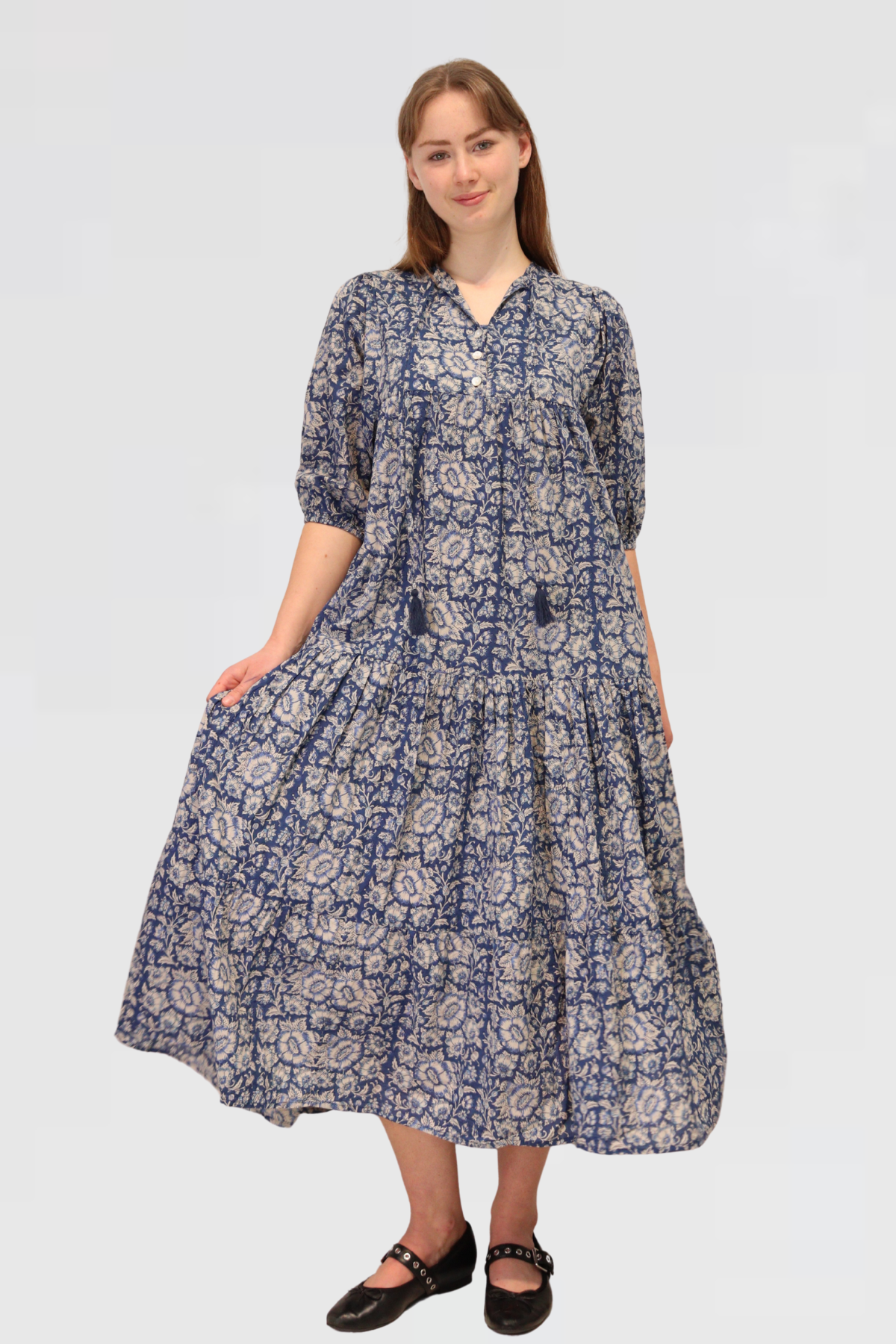 Full front view of model wearing Serene Petals Maxi Dress in blue floral cotton, with V-neckline, tassel ties, and tiered flowy skirt.